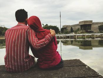 Rear view of couple sitting on retaining wall by lake