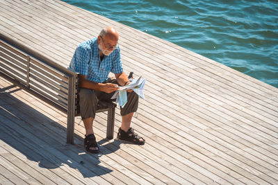 Full length of man reading paper while sitting by lake on bench during sunny day