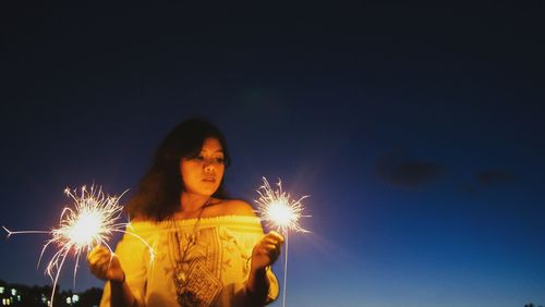 Young woman holding burning sparklers against sky at dusk