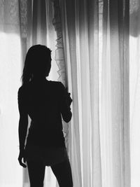 Rear view of woman standing against curtain at window