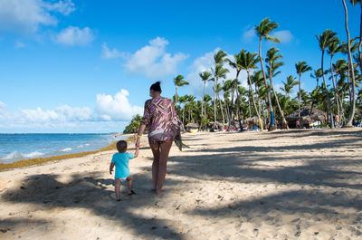 Rear view of mother and son walking at beach against sky