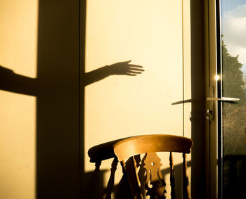 Shadow of a woman's arm and hand above a chair, against a wall next to a glass door in afternoon sun