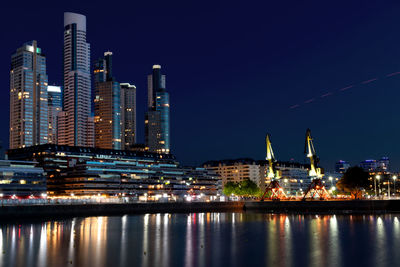 Illuminated modern buildings by river against sky in city