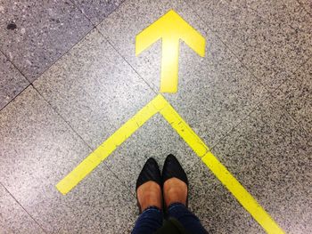 Low section of woman wearing shoes standing by arrow sign on tiled floor