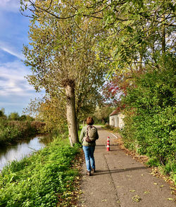 Rear view of woman walking over a footpath in rural area with trees, a farmhouse and a canal