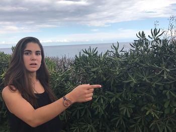 Portrait of woman pointing while standing by plants against cloudy sky