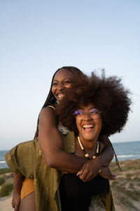 Cheerful young african american female with afro pigtails giving piggyback ride to laughing girlfriend with curly hair while having fun during summer holidays together on seashore