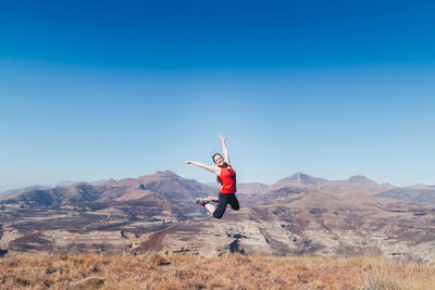 Happy woman with arms raised jumping on mountain against clear sky