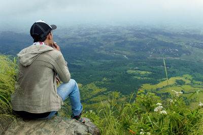 Rear view of man looking at landscape while sitting on mountain against sky