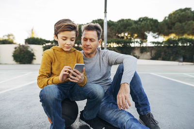 Father and son sitting on basketball outdoor court using cell phone