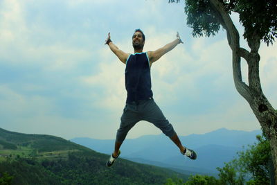 Full length of man with arms raised jumping against landscape and sky