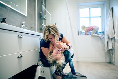 Smiling mother brushing daughter's teeth while girl climbing on window in bathroom