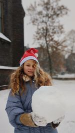 Close up portrait of pretty girl with curly hair in santa's red hat holding a big snowball.