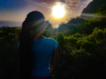 Rear view of woman standing by plants against sun during sunset