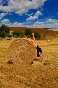 Full length of woman doing handstand by hay bale on agricultural field