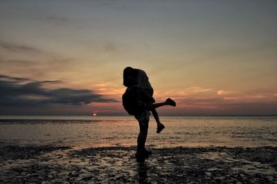 Man lifting woman on wet shore at beach against sky during sunset