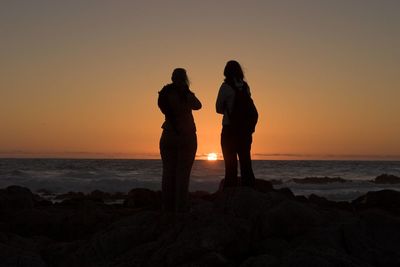 Silhouette people standing on rock at beach against sky during sunset