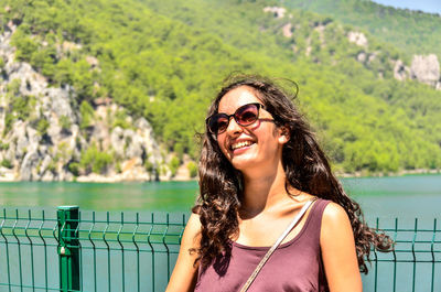 Close-up of smiling young woman in sunglasses standing by railing against lake