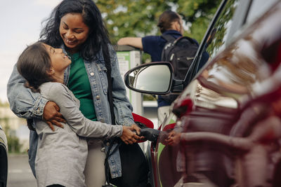 Happy woman embracing daughter while charging electric car at station