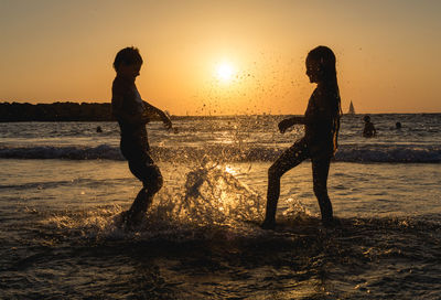 Siblings splashing water in sea against orange sky during sunset