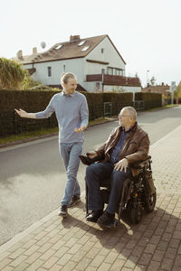 Full length of smiling senior man with disability in motorized wheelchair by young caregiver walking on road during sunn