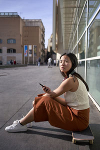 Young woman with smart phone sitting on skateboard during sunny day