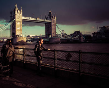 People standing by thames river against tower bridge against sky in city