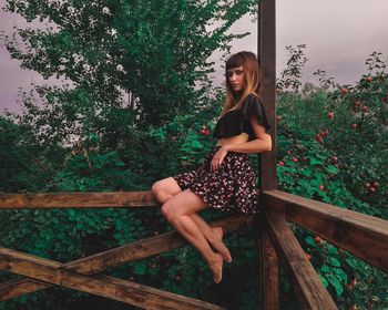Portrait of young woman sitting on wooden railing against trees