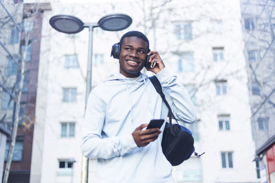 Smiling young man listening to music while using phone outside building