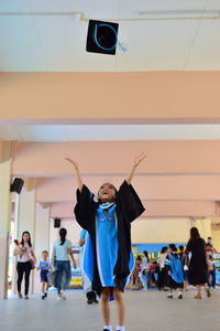 Girl catching mortarboard while standing on floor with people in background