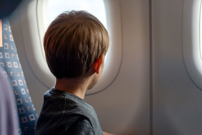 A little boy sits on an airplane and looks out of an illuminator.