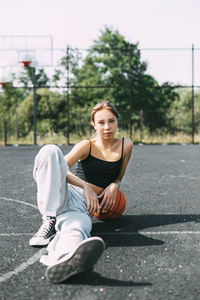 Portrait of a charming girl sitting on a sports field in a park or school with a basketball 