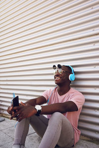 Smiling teenage boy listening music while sitting against wall