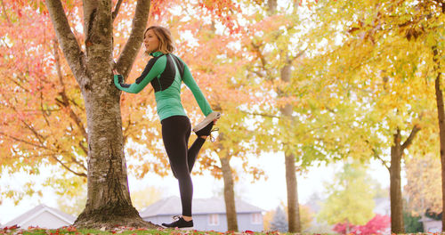 Young woman stretching leg while exercising by tree trunk in park during autumn