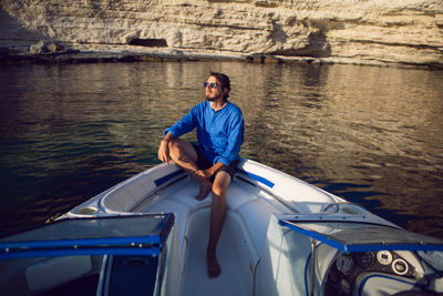 Man with a beard in blue shirt and sunglasses sitting on a yacht near the rocks in the sea in summer