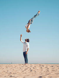 Full body side view of sporty female acrobat performing somersault high in air against blue sky over man standing on sandy seashore