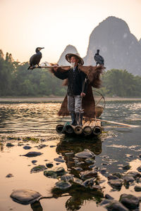 Senior man wearing hat standing on raft against mountain and sky