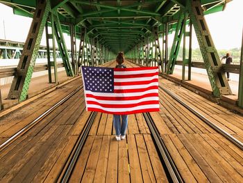 Rear view of woman standing while holding american flag at railway bridge