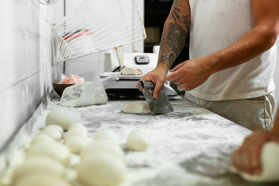 Crop unrecognizable tattooed male baker cutting pieces of raw dough while working together with colleague at floury table in kitchen