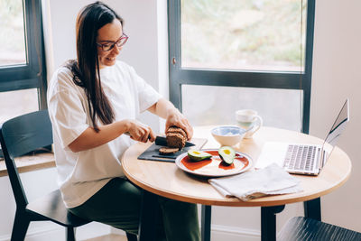 A young woman sitting at a table in front of a laptop makes a healthy breakfast.