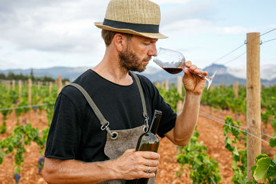 Adult man harvesting ripe grapes from vine on cloudy day on farm serving red wine with bottle in glass and smelling it