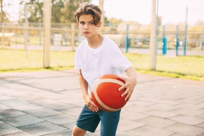 A beautiful teenager in a white t-shirt stands on the basketball court and holds a basketball 