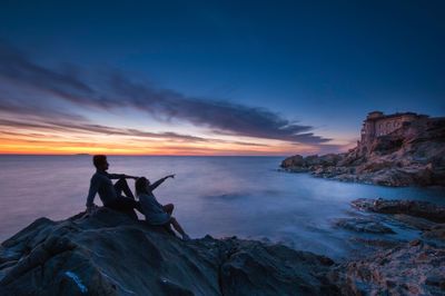 Couple sitting on rock at beach against sky during dusk