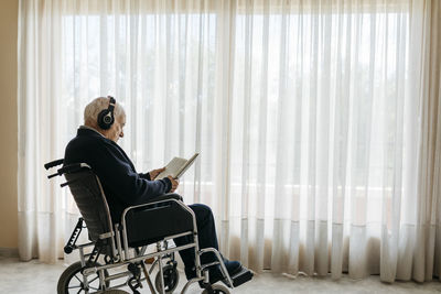 Senior man sitting in wheelchair reading a book while listening music with headphones