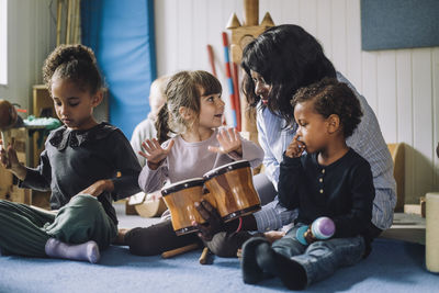 Female teacher teaching girl to play drum kit by classmates in day care center