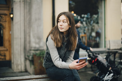 Thoughtful girl with smart phone sitting on bench in city