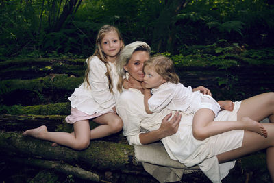 Family blonde mother with two children in a white dress is lying on tree log in the forest barefoot