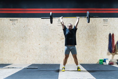 Full body of strong male athlete in sportswear doing clean and jerk with heavy metal barbell during weightlifting training near wall in light gym