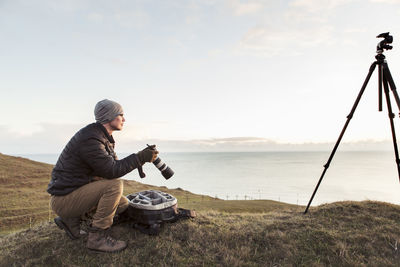 Side view of hiker looking away while holding slr camera on hill by sea against sky