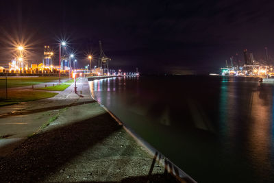 Illuminated street by sea against sky at night